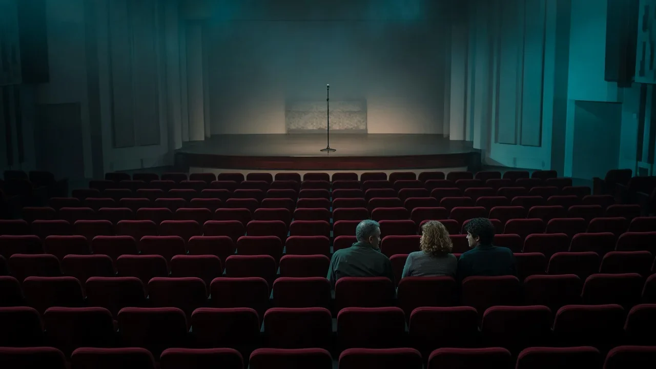 Three people sit in an otherwise empty theater facing a microphone on a bare stage.
